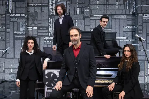 Neri Marcorè, in giacca scura e camicia rossa, siede al centro della scena davanti a un pianoforte; attorno a lui due musiciste e due musicisti vestiti di nero, tra pianoforti e un registratore a bobine. Le pareti sono coperte di pagine di giornale.