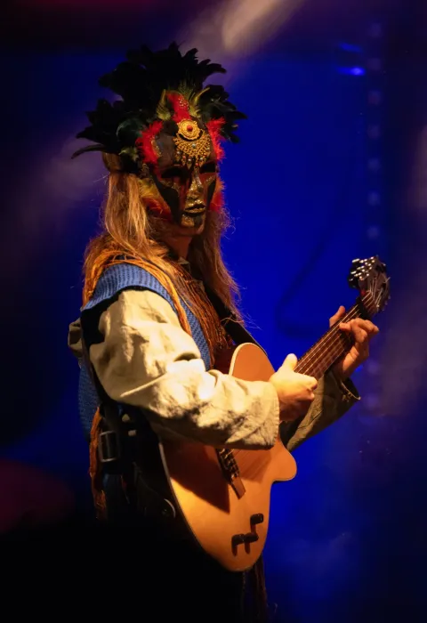 Un musicista degli HAARA suona la chitarra sul palco, con una maschera dorata ornata di piume nere e rosse, una camicia chiara e un gilet blu, davanti a luci blu