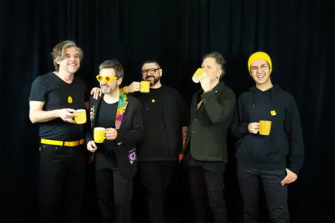 I cinque musicisti degli ALMA Acoustic in piedi davanti a una tenda nera, vestiti di scuro e con dettagli gialli: ognuno tiene una tazza gialla, due bevono, gli altri ridono; uno porta occhiali gialli rotondi, uno un berretto giallo.
