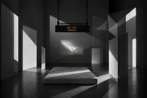 Una sala buia con fasci di luce che entrano di taglio: a terra un materasso bianco, intorno pannelli di vetro e sul fondo uno schermo; dal soffitto pende un cartello luminoso con la scritta «ACT III Operator».