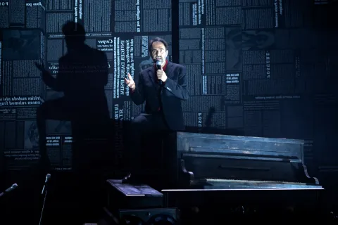 Neri Marcorè canta al microfono seduto sopra un vecchio pianoforte, su un palco in penombra. Alle sue spalle le pareti sono coperte di pagine di giornale e la sua ombra si allunga sulla sinistra.