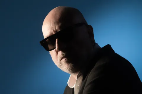 Ritratto di Mario Biondi su sfondo blu: calvo, con la barba bianca corta, occhiali da sole scuri, camicia bianca e giacca scura, il viso in parte in ombra.