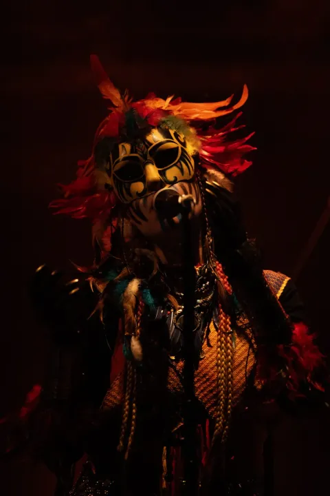 La voce degli HAARA canta al microfono con una maschera gialla dipinta, piume rosse e arancioni sulla testa e un costume intrecciato, in penombra su sfondo scuro