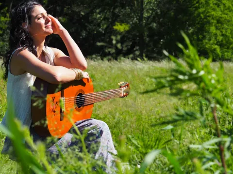 Lia Seddio seduta nell'erba al sole, con una chitarra classica arancione appoggiata in grembo: tiene una mano sulla fronte e sorride a occhi socchiusi, sullo sfondo un prato e alberi.