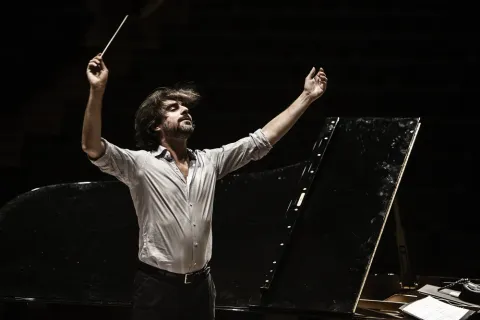 Alessandro Cadario dirige con le braccia alzate e la bacchetta nella mano destra, in camicia chiara, accanto a un pianoforte a coda su sfondo scuro.