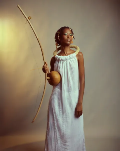 Una musicista con un abito bianco lungo e un collare intrecciato tiene in mano un berimbau, arco musicale con una zucca come cassa, su sfondo chiaro.