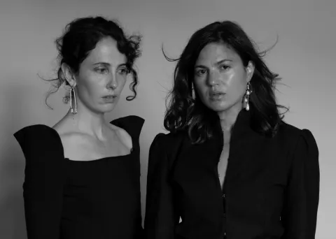 Ritratto in bianco e nero di Ania Catherine e Dejha Ti, il duo Operator: due donne vestite di nero davanti a un fondo grigio, una con i capelli ricci raccolti e un grande orecchino a spilla da balia, l'altra con i capelli mossi e un orecchino di perle.