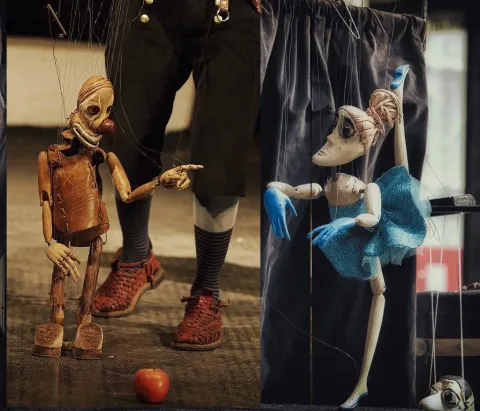 Due marionette a fili di legno, un clown con il naso rosso e una ballerina con il tutù azzurro, mosse dal marionettista di cui si vedono solo le gambe; a terra, una mela.